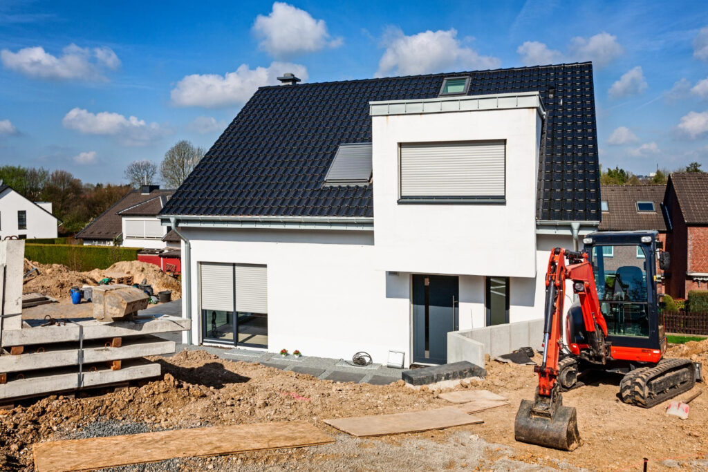 A new family home that is white with a black roof. A red digger sits to the side of the house to show that the building work goes on.